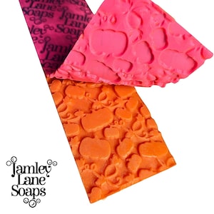 May include: Two rectangular silicone texture mats for soap making. One mat is bright orange with a raised pumpkin pattern, and the other is vibrant pink with a repeating Jamley Lane Soaps logo. The logo text is clearly visible on the pink mat.