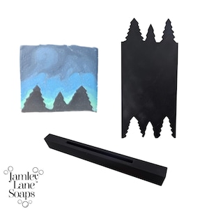 May include: A black silicone soap mold with a design of three pine trees. The mold is used to create a soap bar with a mountain scene. The soap bar is blue and green with a white sky.