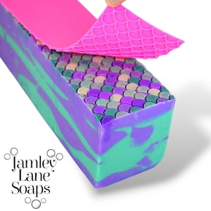 Puede incluir: Una barra de jabón con un diseño de escamas de sirena. El jabón es una mezcla de morado, verde y blanco. Un molde de silicona rosa se está despegando del jabón.