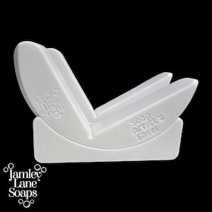Op de afbeelding: Een witte plastic zeepkunstenaar's ezel met de tekst "Soap Artist's Easel" en "Jamley Lane Soaps" erop gedrukt. De ezel heeft een gebogen basis en schuine steunen.