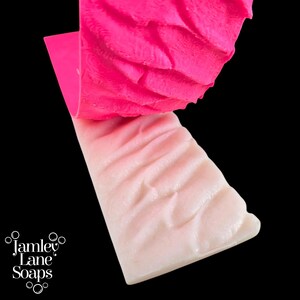 May include: A pink silicone mold with a wavy texture is being peeled away from a white soap bar with a similar wavy texture. The soap bar has a subtle pink gradient.