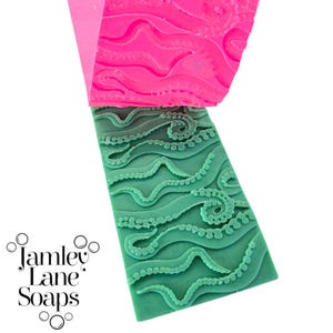 Könnte beinhalten: Ein grünes Seifenstück mit einem Oktopus-Tentakel-Design, neben einer rosa Form mit dem gleichen Muster. Das Seifenstück hat eine rechteckige Form und die Tentakel sind erhaben. Das Logo "Jamley Lane Soaps" ist unten links zu sehen.