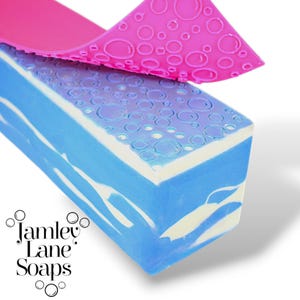 Può includere: Una saponetta blu e bianca con un design a bolle sulla parte superiore. Il sapone viene rimosso da uno stampo in silicone rosa con un design a bolle.
