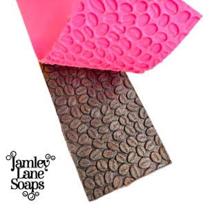 May include: A brown soap bar with a coffee bean pattern is partially revealed beneath a bright pink mold. The soap bar has a textured surface with the coffee bean design. The Jamley Lane Soaps logo is visible in the lower left corner.
