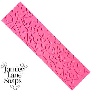 May include: A bright pink soap mold with a repeating leaf and vine pattern. The rectangular mold is angled, showcasing the detailed design. The words "Jamley Lane Soaps" are printed in black script on the lower left.