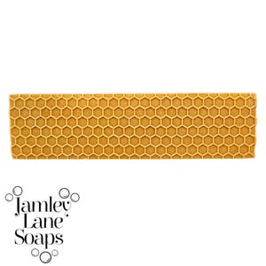 May include: A golden-yellow soap bar with a honeycomb pattern. The rectangular bar features a detailed, repeating hexagonal design, evoking a natural aesthetic. The logo "Jamley Lane Soaps" is visible at the bottom of the image.