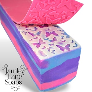 May include: A pink silicone stamp with a butterfly design is being used to create a pattern on a multi-colored soap bar. The soap bar has layers of pink, blue, and purple. The soap bar is labeled "Jamley Lane Soaps".