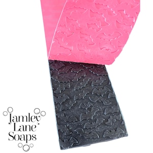 May include: A close-up of two soap bars. One is black with a bat pattern, and the other is pink. The logo "Jamley Lane Soaps" is in the lower left corner. The soap bars are likely handmade and suitable for Halloween.