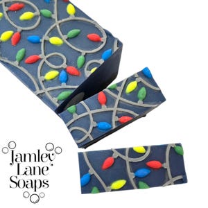 May include: Dark blue soap bars decorated with a string of colorful Christmas lights. The lights are red, green, yellow, and blue. The soap bars have a gray outline of the lights. The brand name "Jamley Lane Soaps" is in the lower left corner.