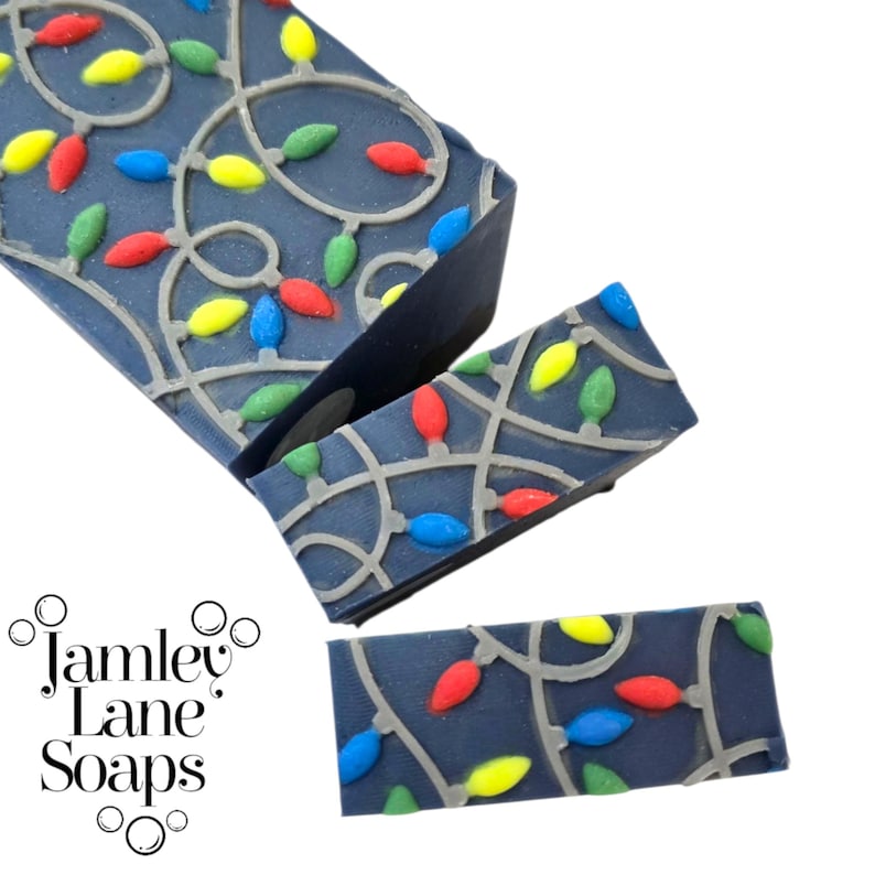 May include: Dark blue soap bars decorated with a string of colorful Christmas lights. The lights are red, green, yellow, and blue. The soap bars have a gray outline of the lights. The brand name "Jamley Lane Soaps" is in the lower left corner.