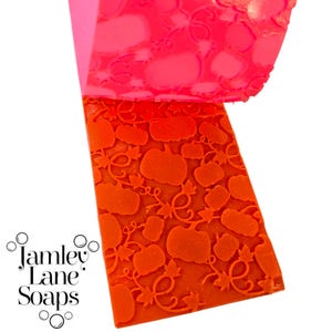 May include: A bright orange soap mold with a pumpkin and vine pattern. The mold is made of flexible silicone and is partially peeled back to reveal the embossed design. The brand name "Jamley Lane Soaps" is printed in white.