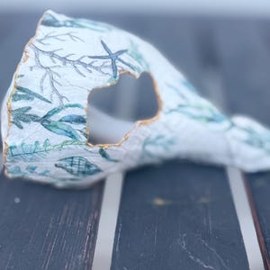 Könnte beinhalten: Eine dekorative, abstrakte Skulptur mit einem ausgeschnittenen Mittelteil. Die weiße Oberfläche ist mit einem Meeresmuster mit blau-grünen Algen, Muscheln und einem Seestern verziert. Die Kanten sind goldfarben umrandet und schaffen ein einzigartiges Kunstwerk.
