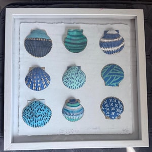 May include: A framed artwork featuring nine painted seashells in shades of blue, teal, and gold. The shells are arranged on a textured white background within a white wooden frame. The artwork has a coastal theme.