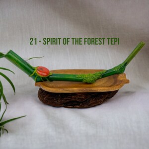 May include: A green and brown wooden pipe with a red stone accent. The pipe is resting in a wooden bowl on a white surface. The text "21 - SPIRIT OF THE FOREST TEPI" is visible in the image.
