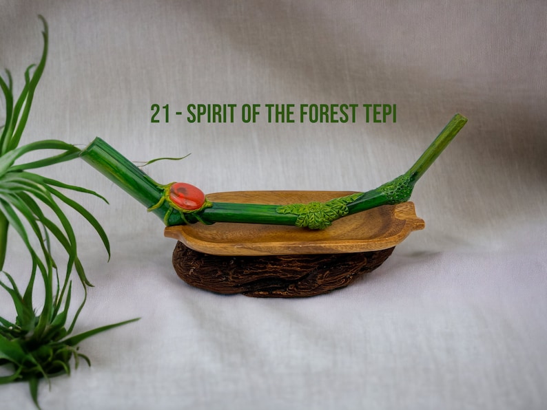 May include: A green and brown wooden pipe with a red stone accent. The pipe is resting in a wooden bowl on a white surface. The text "21 - SPIRIT OF THE FOREST TEPI" is visible in the image.