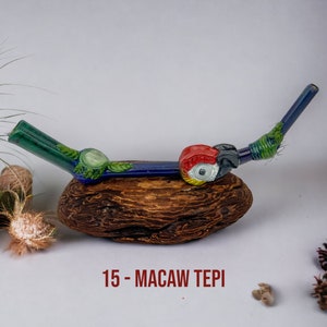 May include: A blue and green glass pipe with a macaw design. The pipe is resting on a piece of driftwood. 15 - MACAW TEPI