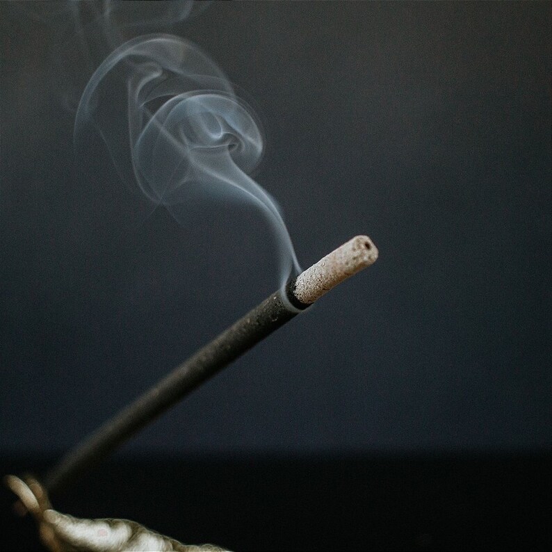 May include: A black incense stick with a brown tip is burning, releasing a wisp of white smoke against a dark gray background.