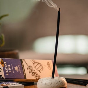 May include: A white ceramic incense holder with a black incense stick burning and smoke rising. The incense stick is labeled "ALFAZEMA E MEL" with the brand name "nirvana" printed on the box.