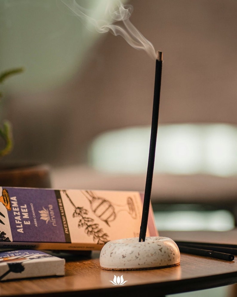 May include: A white ceramic incense holder with a black incense stick burning and smoke rising. The incense stick is labeled "ALFAZEMA E MEL" with the brand name "nirvana" printed on the box.