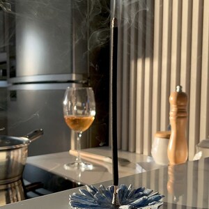 May include: A black incense stick burning in a blue ceramic incense holder. Smoke rises from the stick. The holder is shaped like a flower.