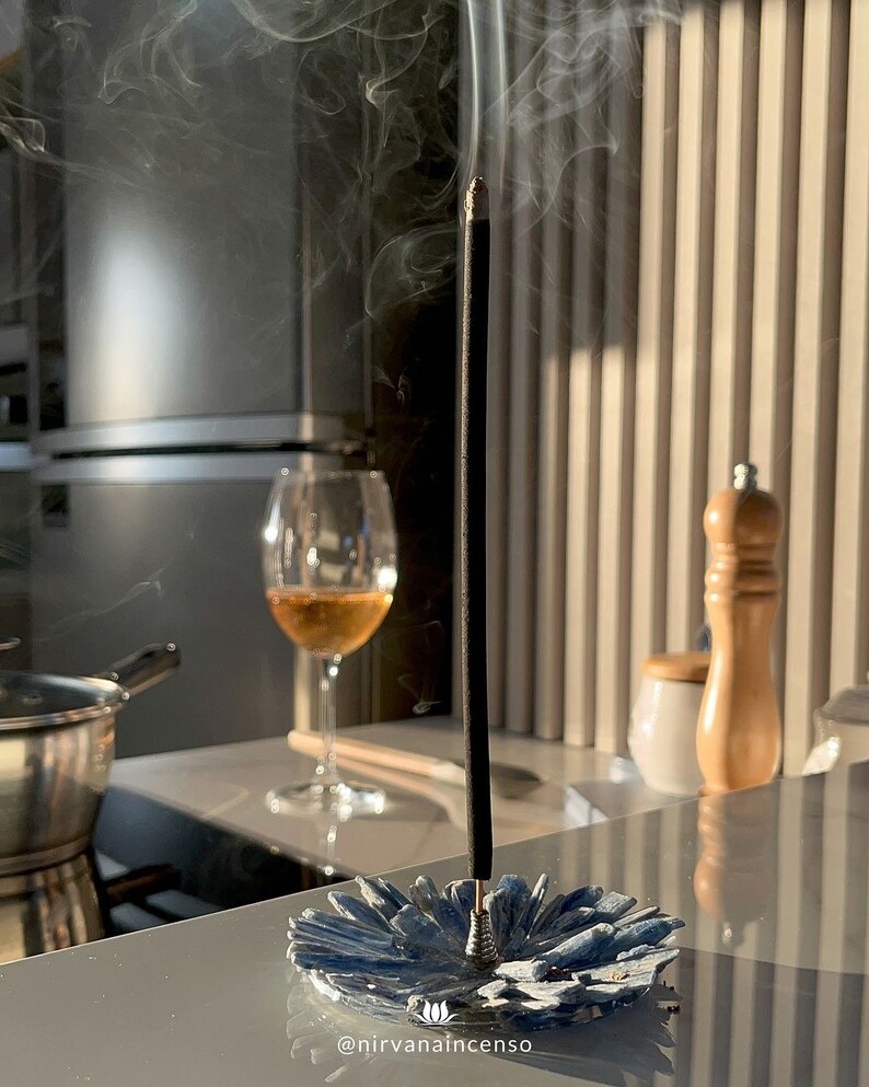 May include: A black incense stick burning in a blue ceramic incense holder. Smoke rises from the stick. The holder is shaped like a flower.