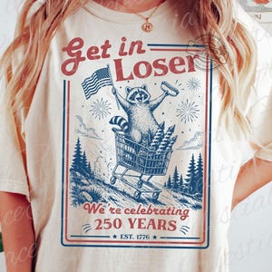 May include: Ivory-colored t-shirt with a graphic featuring a raccoon pushing a shopping cart filled with fireworks. The raccoon holds an American flag and a hot dog. Text reads "Get in Loser" and "We're celebrating 250 YEARS EST. 1776."