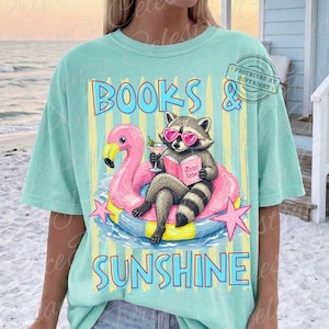 May include: A light teal t-shirt with a graphic of a raccoon wearing heart-shaped sunglasses, reading a book while floating on a pink flamingo pool float. The text "BOOKS & SUNSHINE" is above and below the graphic.