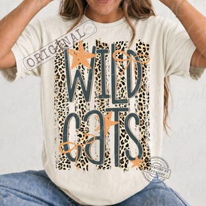 May include: A cream-colored short-sleeve t-shirt featuring a graphic design with vertical leopard print stripes. The text "WILD CATS" is printed in dark gray letters, accented with light orange star shapes and thin, hand-drawn style line art bows.