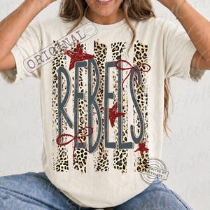 May include: A cream-colored short-sleeve t-shirt featuring a graphic design with vertical leopard print stripes. The word REBELS is printed in large dark gray letters over the stripes, accented by red stars and thin red line art flourishes.
