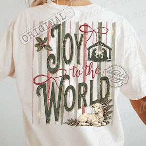 May include: A cream-colored t-shirt featuring a back graphic with the text Joy to the World in dark green and red. The design includes a nativity scene, a red bow, holly leaves, and a lamb resting among pine branches over vertical stripes.