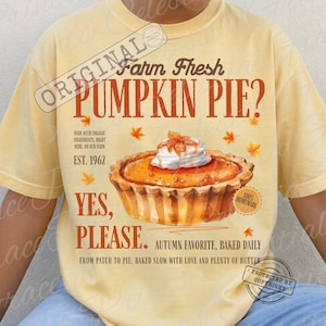 May include: A pale yellow t-shirt featuring a graphic of a pumpkin pie with whipped cream. The text reads: Farm Fresh PUMPKIN PIE? YES, PLEASE. MADE WITH ORGANIC INGREDIENTS, RIGHT HERE ON OUR FARM EST. 1962. AUTUMN FAVORITE, BAKED DAILY.