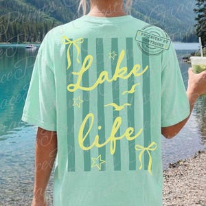 Puede incluir: Camiseta verde claro con la frase "Lake Life" en amarillo, adornada con estrellas, lazos y p&aacute;jaros. La camiseta tiene rayas verticales y est&aacute; etiquetada como "Comfort Colors Island Reef."
