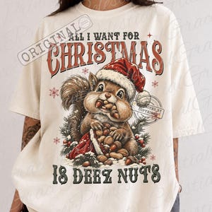 May include: A cream-colored t-shirt featuring a graphic of a squirrel wearing a Santa hat, holding acorns. The text reads ALL I WANT FOR CHRISTMAS IS DEEZ NUTS. The design includes red and green accents, pine branches, and small snowflakes.