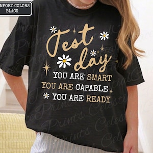 May include: Black t-shirt with the words "Test Day" in a cursive font, decorated with daisies and stars. The shirt also features the phrases "You are smart, you are capable, you are ready."