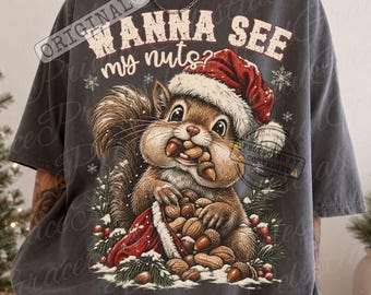 May include: A charcoal grey t-shirt featuring a graphic of a squirrel wearing a red and white Santa hat. The squirrel holds acorns in its mouth and paws, surrounded by pine branches and berries. The text reads, "WANNA SEE my nuts?"