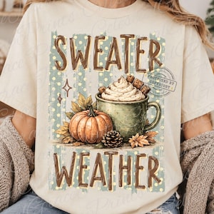 Op de afbeelding: Een crèmekleurig t-shirt met een grafische print met de tekst "SWEATER WEATHER" in bruine letters. Het ontwerp bevat een groene mok gevuld met slagroom, een oranje pompoen, dennenappels en herfstbladeren op een achtergrond met stippen.