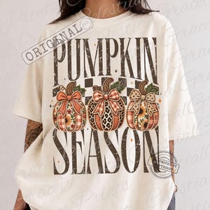 May include: An off-white short-sleeve t-shirt featuring the text PUMPKIN SEASON in dark brown block letters. Between the words are three illustrated pumpkins with patterned surfaces, each adorned with a decorative bow in a matching print.
