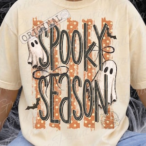 May include: A cream-colored t-shirt featuring a graphic print with the text "SPOOKY SEASON" in black outlined letters. The design includes two white ghosts, orange vertical stripes with white polka dots, and small black bat silhouettes.