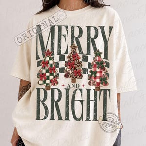 May include: An off-white short-sleeve t-shirt featuring the text MERRY AND BRIGHT in dark green. Between the words are three stylized Christmas trees: one with a green checkered pattern, one with a leopard print, and one with a red plaid pattern.