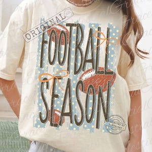 Op de afbeelding: Een crèmekleurig t-shirt met een grafische print met de tekst FOOTBALL SEASON in grote bruine letters. Het ontwerp bevat illustraties van drie bruine footballs, lichtblauwe verticale strepen en oranje lintaccenten.