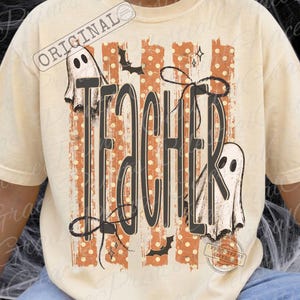 May include: A cream-colored t-shirt featuring a graphic design with the word TEACHER in large black letters. The background of the design includes orange vertical stripes with white polka dots, two white ghosts, black bats, and stars.