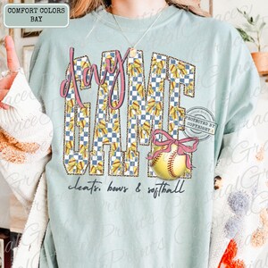 May include: A light blue t-shirt with the words "Day Game" in a pink, checkered, and bow-filled design. A softball with a pink bow is featured below the text. The words "cleats, bows & softball" are written at the bottom.