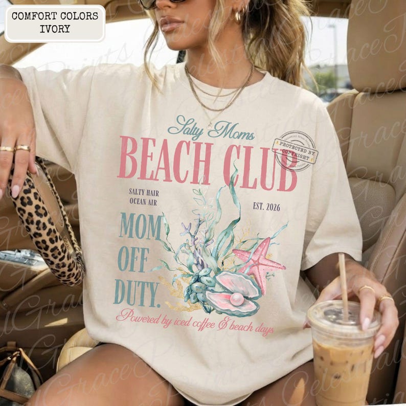 Puede incluir: Camiseta de color marfil con el texto "Salty Moms Beach Club" en rosa y azul. El dise&ntilde;o incluye una estrella de mar, conchas marinas y las palabras "Mom Off Duty". La camiseta tambi&eacute;n tiene el texto "Powered by iced coffee & beach days."