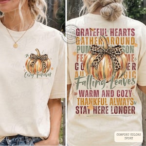May include: Ivory-colored t-shirt featuring a pumpkin design with a leopard print bow. The front of the shirt has the text "Cozy Forever." The back of the shirt has a larger pumpkin design and the phrases "Grateful Hearts," "Falling Leaves," and "Warm and Cozy."