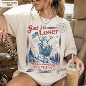 May include: Ivory-colored t-shirt with a graphic featuring a raccoon in a shopping cart, holding an American flag and a hot dog. Text reads "Get in Loser" and "We're celebrating 250 YEARS EST. 1776".