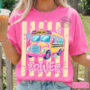 May include: A bright pink t-shirt featuring a graphic of a yellow school bus with the text SCHOOL BUS DRIVER. The design includes light yellow vertical stripes and light blue and green star accents. The shirt has a crew neckline and short sleeves.