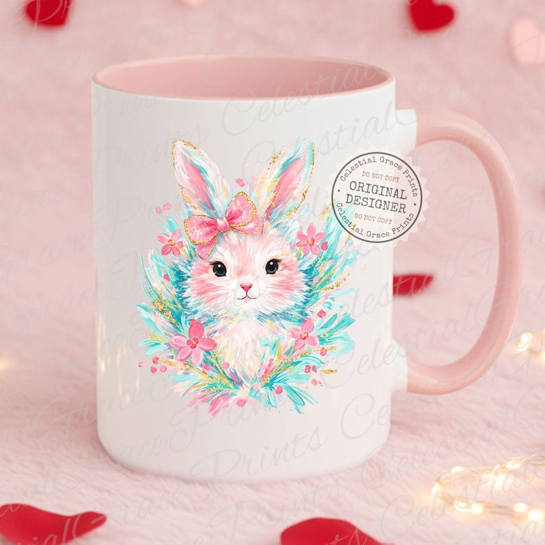 May include: White ceramic mug with a pink interior and handle. The mug features a watercolor illustration of a pink bunny with a pink bow, surrounded by flowers and greenery. The mug has the text "ORIGINAL DESIGNER" on it.