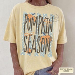 May include: A pale yellow short-sleeve t-shirt featuring a graphic print with the text PUMPKIN SEASON in dark brown letters. The design includes light blue vertical stripes with white polka dots and orange pumpkin illustrations with thin bows.