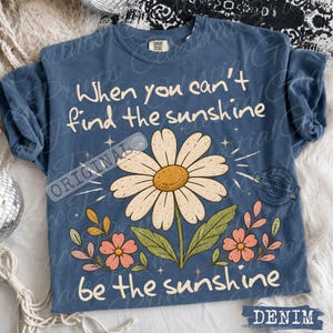 May include: A blue short-sleeved t-shirt featuring a large white daisy graphic with a yellow center, surrounded by smaller pink and orange flowers. The text reads, When you can't find the sunshine, be the sunshine, in a cream-colored script font.