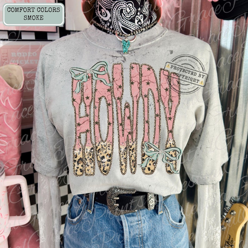 Peut inclure: Un t-shirt gris &agrave; manches longues avec le mot "HOWDY" en rose, imprim&eacute; l&eacute;opard et des &eacute;toiles. Le t-shirt pr&eacute;sente des n&oelig;uds bleu clair et est associ&eacute; &agrave; un jean bleu et une ceinture noire avec une boucle argent&eacute;e. Le texte "COMFORT COLORS SMOKE" est en haut &agrave; gauche.